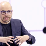 Man in glasses speaking, gesturing with hands, wearing a dark suit jacket with a microphone headset against a geometric ba...