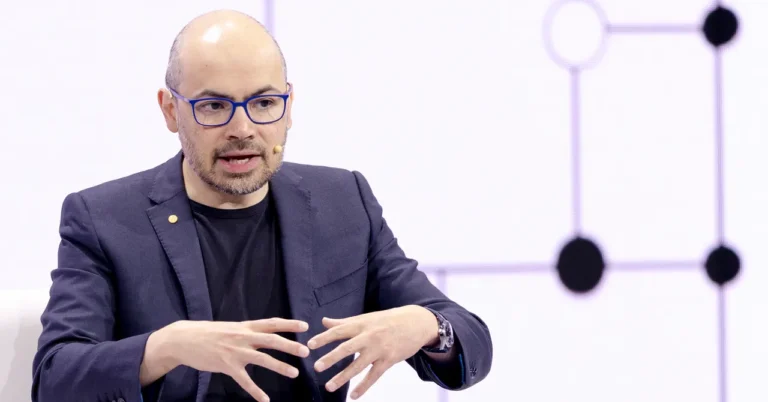 Man in glasses speaking, gesturing with hands, wearing a dark suit jacket with a microphone headset against a geometric ba...