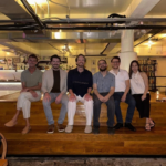 Seven people sitting on wooden steps in an industrial-style room with exposed pipes and bookshelves in the background.