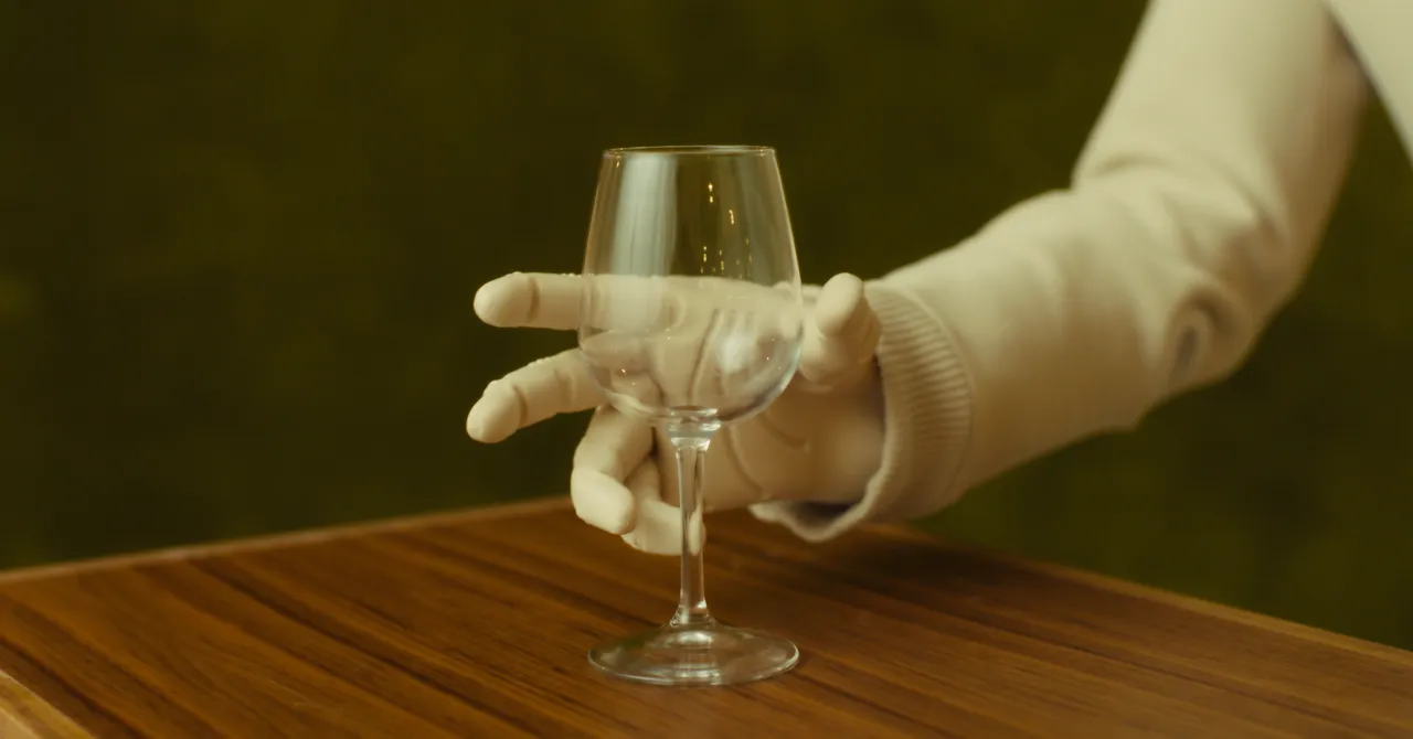 Robot hand with five fingers holding a wine glass on a wooden table, against a blurred green background.