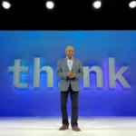 Man in a suit stands on stage with blue "think" backdrop, under bright overhead lights.