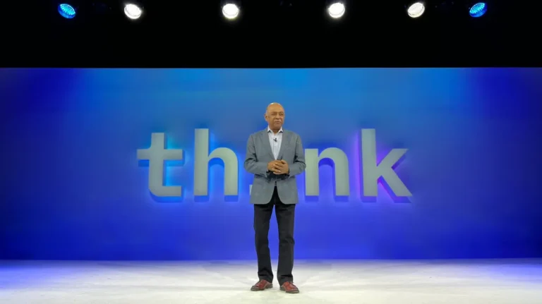 Man in a suit stands on stage with blue "think" backdrop, under bright overhead lights.