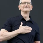 Man in black shirt with hand on chest, smiling, against a dark background.