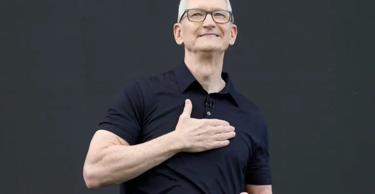 Man in black shirt with hand on chest, smiling, against a dark background.