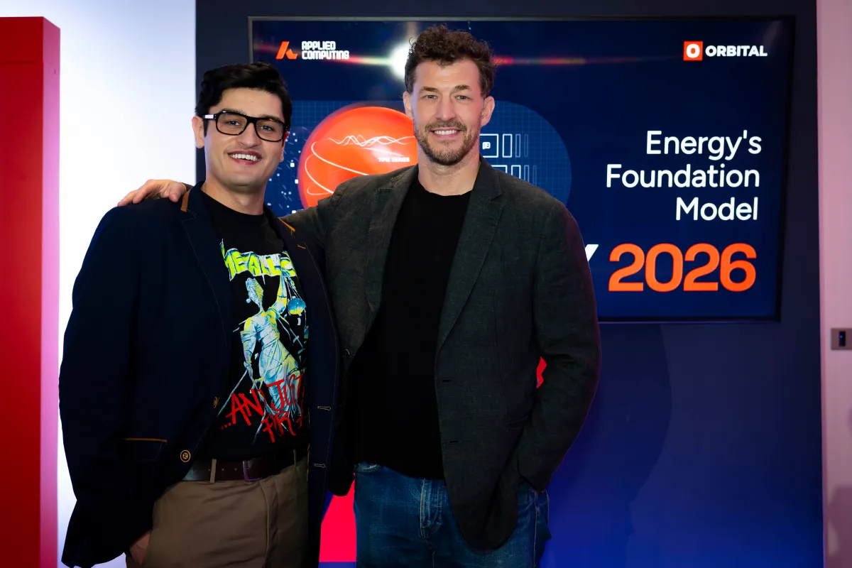 Two people smiling in front of a screen displaying "Energy's Foundation Model" and "2026" with logos for Applied Computing...