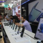 Customers in an electronics store examining smartphones on display, large screen showing Galaxy Z Fold7, promotional poste...