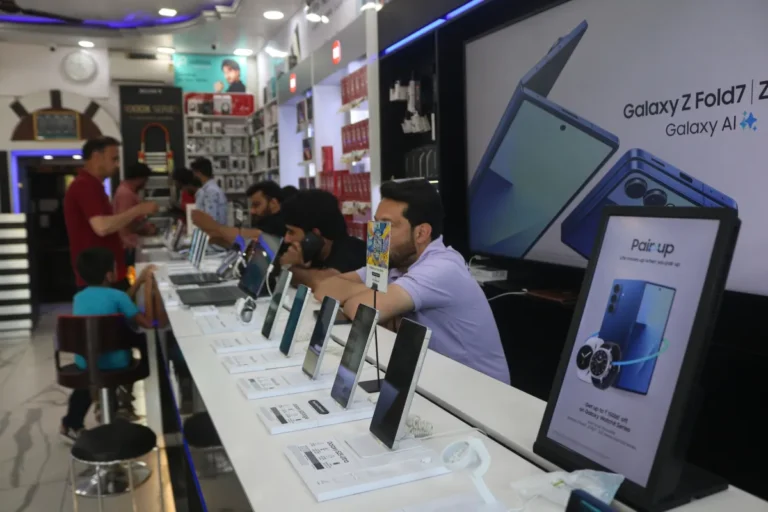 Customers in an electronics store examining smartphones on display, large screen showing Galaxy Z Fold7, promotional poste...