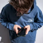 Person in a blue hoodie looking down at a smartphone, standing against a light gray wall.