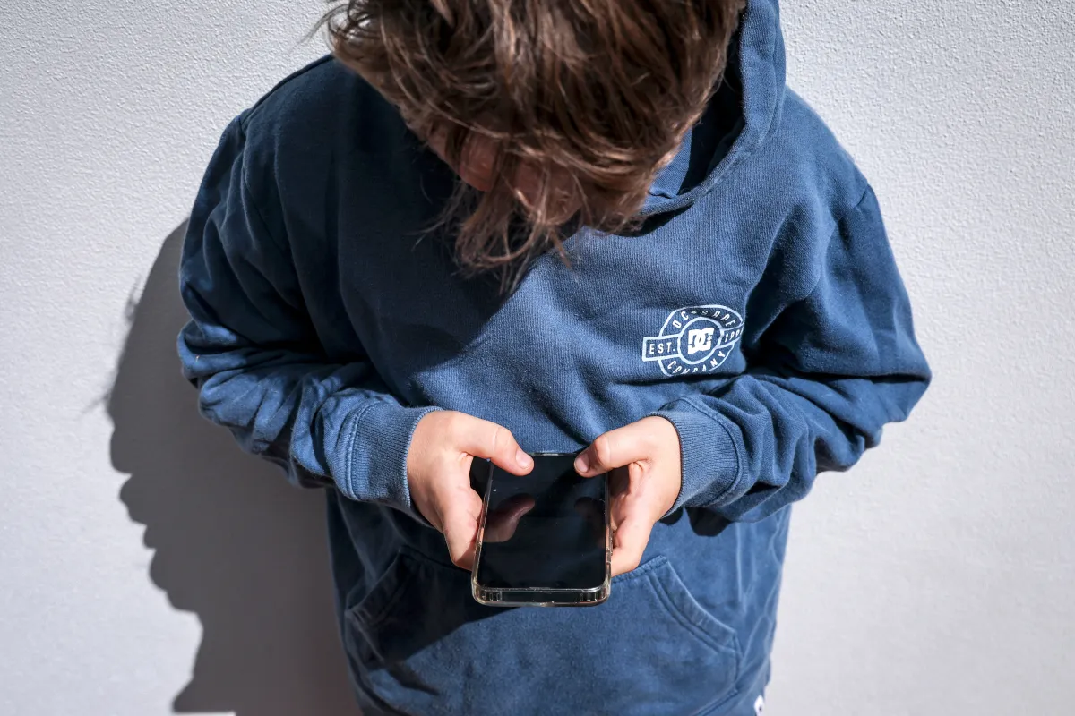 Person in a blue hoodie looking down at a smartphone, standing against a light gray wall.