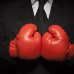 Person in a suit wearing red boxing gloves, arms crossed in front of chest, against a dark background.