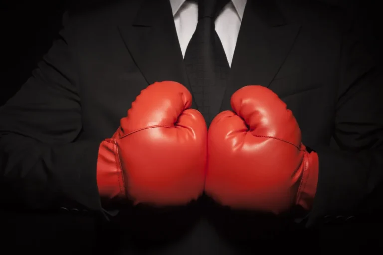 Person in a suit wearing red boxing gloves, arms crossed in front of chest, against a dark background.