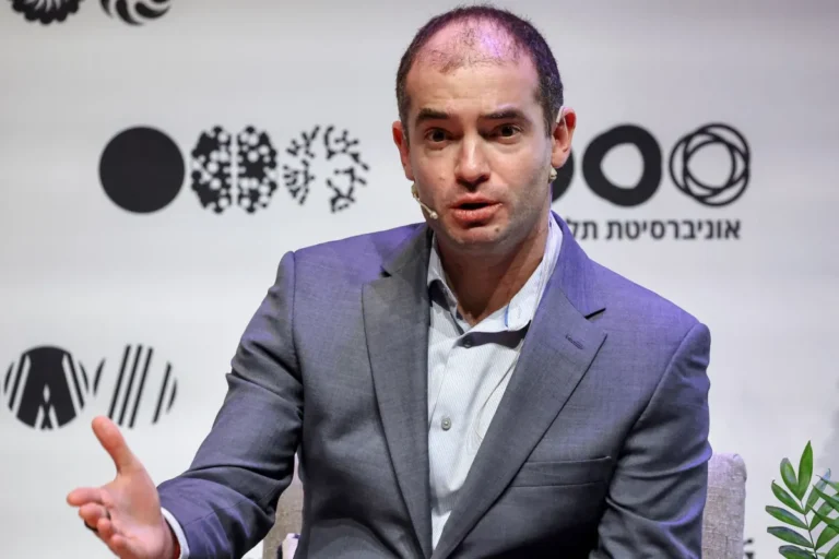 Man in a suit speaks on stage with a headset mic, abstract graphics and text in background, plant in corner.