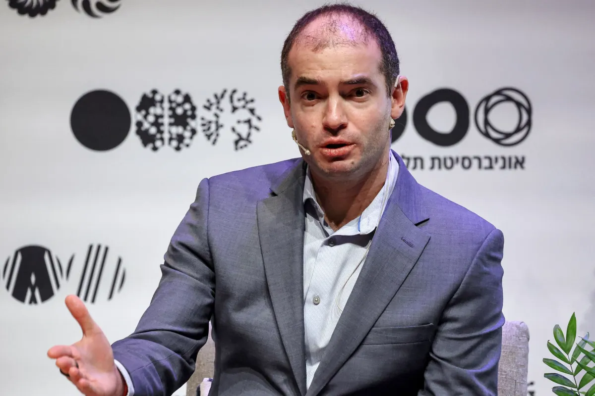 Man in a suit speaks on stage with a headset mic, abstract graphics and text in background, plant in corner.