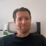 Man in black shirt with earbuds sits in a modern office, neon smiley face light and white robot figures in the background.