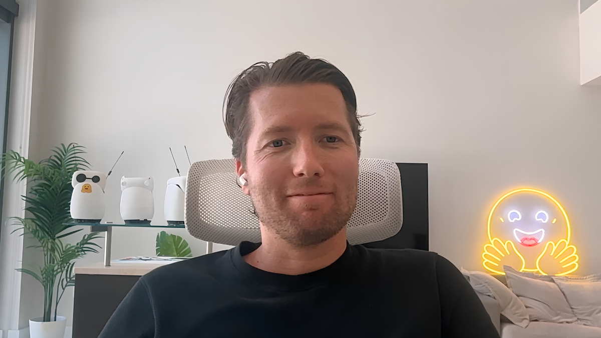 Man in black shirt with earbuds sits in a modern office, neon smiley face light and white robot figures in the background.