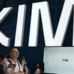 People holding phones stand under large "KIMI" signage, with a screen also displaying "KIMI" in an indoor setting.