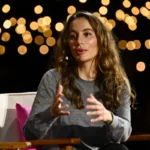 Person speaking on stage, wearing a gray sweater, with bokeh lights in the background and seated on a white chair.