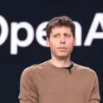 Person in a brown shirt stands in front of a dark background with large, partially visible text "OpenAI".