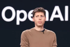 Person in a brown shirt stands in front of a dark background with large, partially visible text "OpenAI".