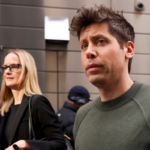 Two individuals walking outdoors, one in a green sweater, the other in a black jacket holding a phone, with a building in...