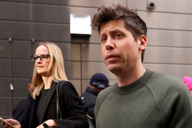 Two individuals walking outdoors, one in a green sweater, the other in a black jacket holding a phone, with a building in...