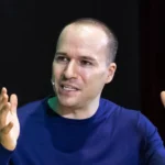 Man in blue shirt speaking with hands raised, wearing a headset microphone, dark background.