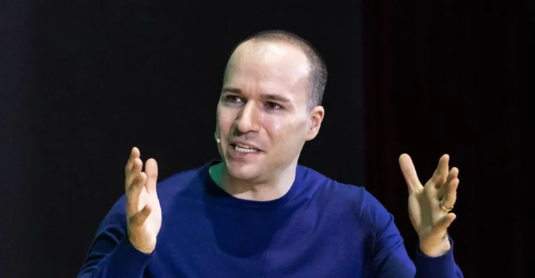 Man in blue shirt speaking with hands raised, wearing a headset microphone, dark background.