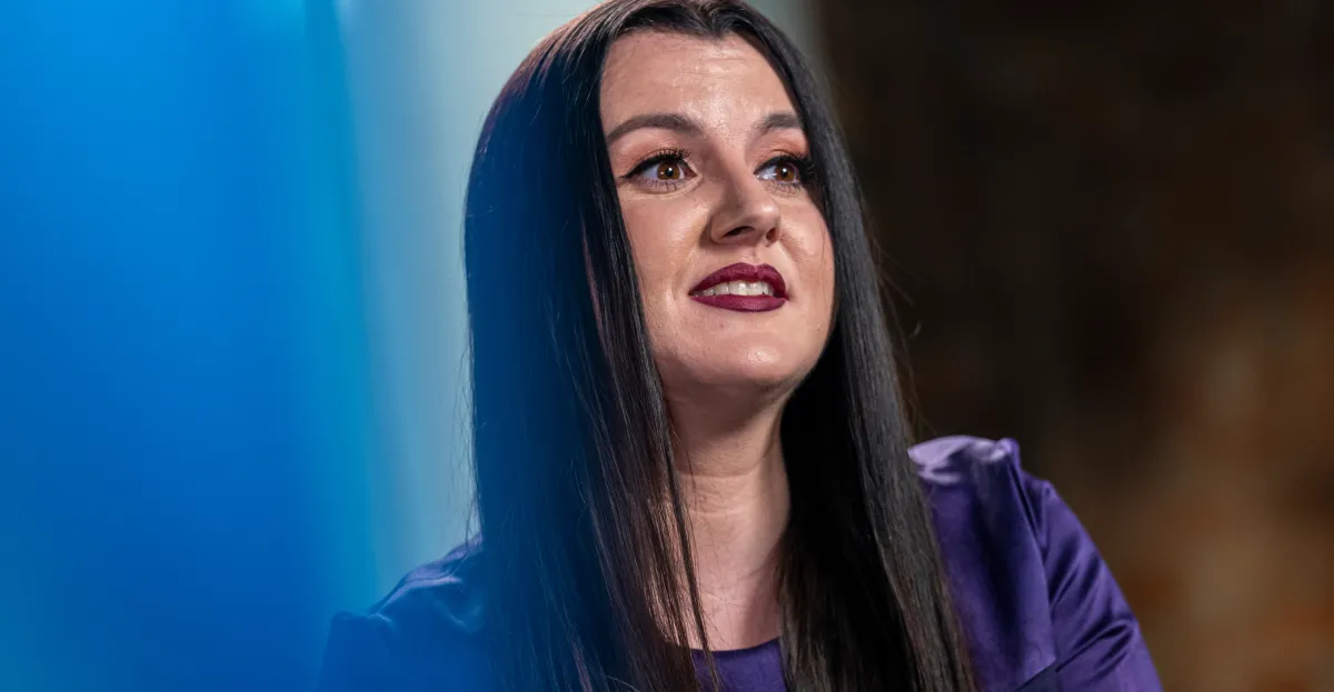Person with long dark hair and purple lipstick, wearing a purple top, looking upwards against a blurred background.