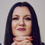 Person with long dark hair, wearing purple lipstick, resting chin on clasped hands with red painted nails, neutral backgro...
