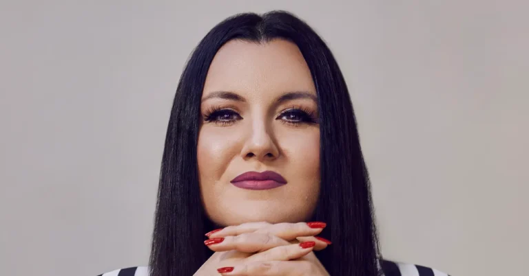 Person with long dark hair, wearing purple lipstick, resting chin on clasped hands with red painted nails, neutral backgro...