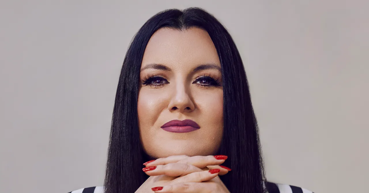 Person with long dark hair, wearing purple lipstick, resting chin on clasped hands with red painted nails, neutral backgro...