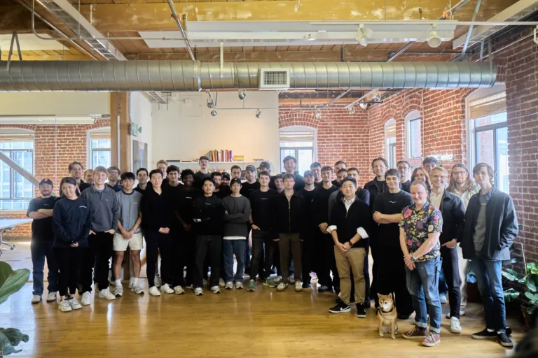 Group of people posing indoors in a brick-walled room with wooden floors; a dog stands in the front.