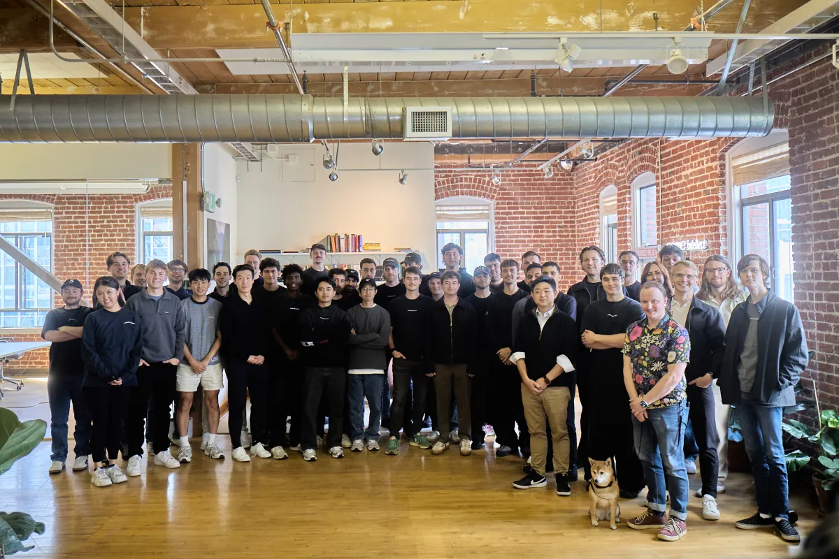 Group of people posing indoors in a brick-walled room with wooden floors; a dog stands in the front.