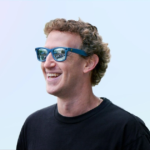 Smiling person wearing blue sunglasses and a black shirt against a light blue background.