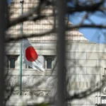 Japanese flag on a pole in front of a stone building with decorative carvings, viewed through blurred branches.