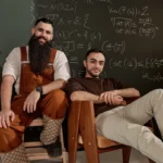 Two men sitting in front of a chalkboard with mathematical equations, one wearing overalls and boots, the other in casual...