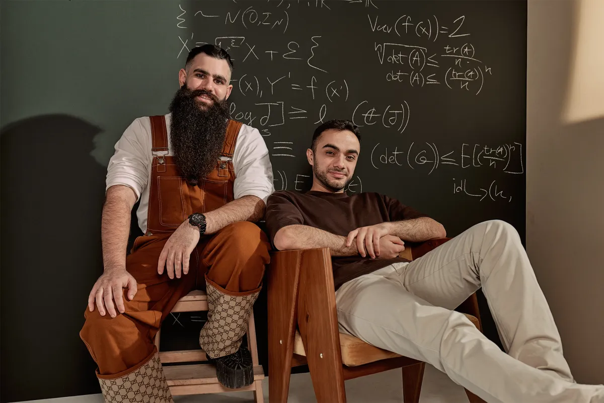 Two men sitting in front of a chalkboard with mathematical equations, one wearing overalls and boots, the other in casual...