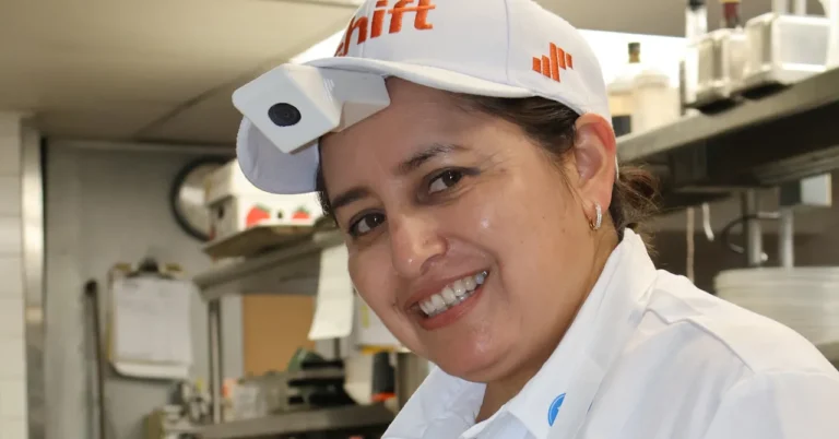 Person in a kitchen wearing a white cap with a camera attached, smiling, shelves and kitchen items in the background.