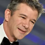 Man in a tuxedo smiling at an event with a blurred blue and gray striped background.
