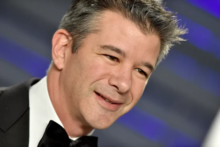 Man in a tuxedo smiling at an event with a blurred blue and gray striped background.