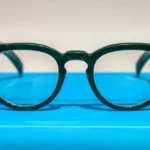 Green smart glasses with round lenses and small cameras on each side, displayed on a bright blue surface.