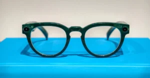 Green smart glasses with round lenses and small cameras on each side, displayed on a bright blue surface.