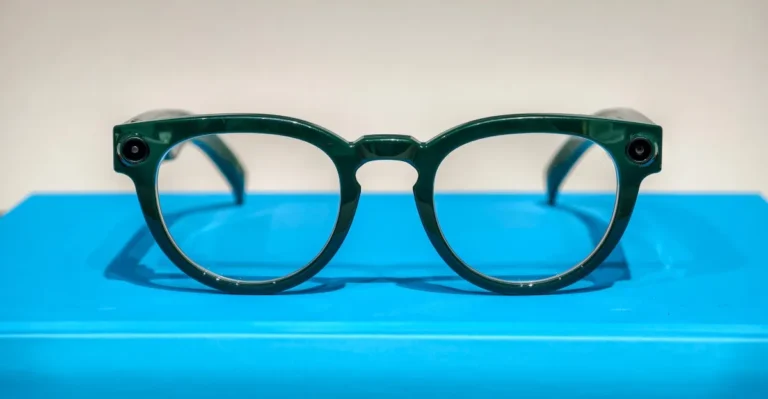 Green smart glasses with round lenses and small cameras on each side, displayed on a bright blue surface.