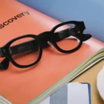 Black smart glasses on an orange booklet labeled "BrandDiscovery," with a cup of coffee and papers nearby.