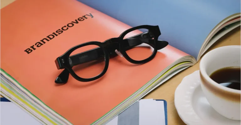 Black smart glasses on an orange booklet labeled "BrandDiscovery," with a cup of coffee and papers nearby.