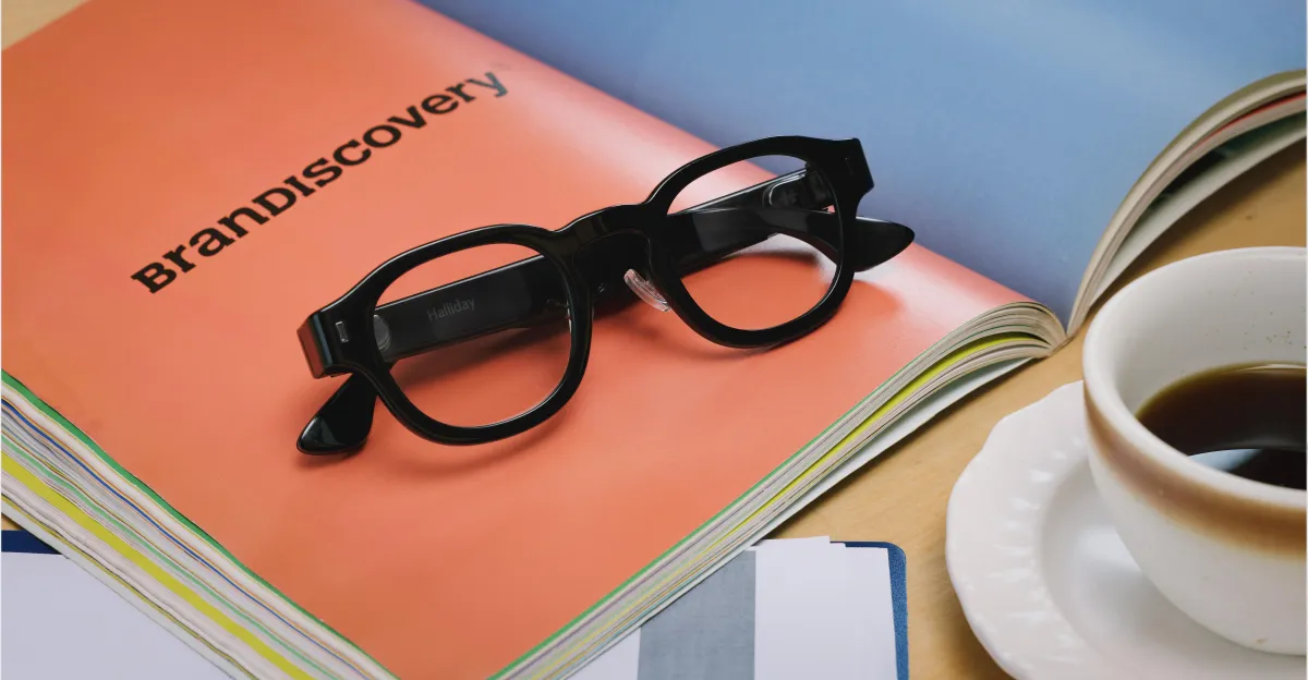 Black smart glasses on an orange booklet labeled "BrandDiscovery," with a cup of coffee and papers nearby.