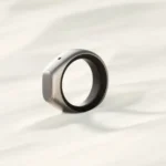 A sleek silver smart ring with a small button rests on a light, textured surface.