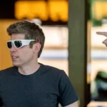 Man wearing smart glasses, looking left, with another person's hand pointing in the background.