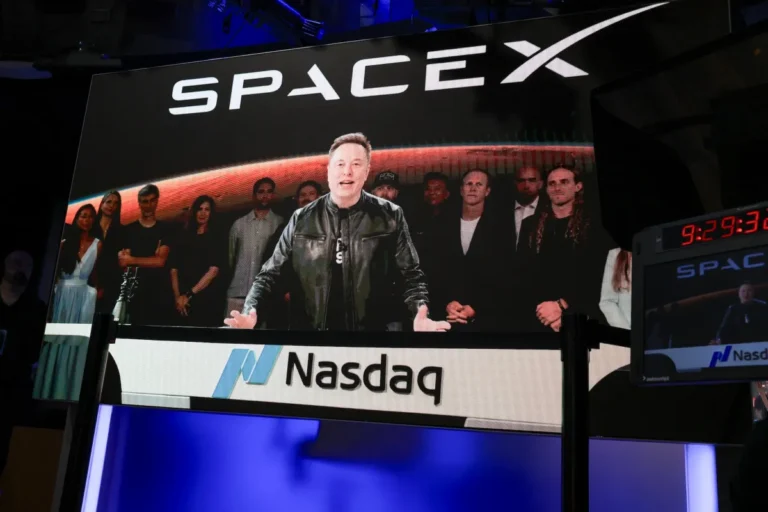 A large screen displays a man speaking with a group behind him, SpaceX logo above, Nasdaq sign below, digital clock shows...