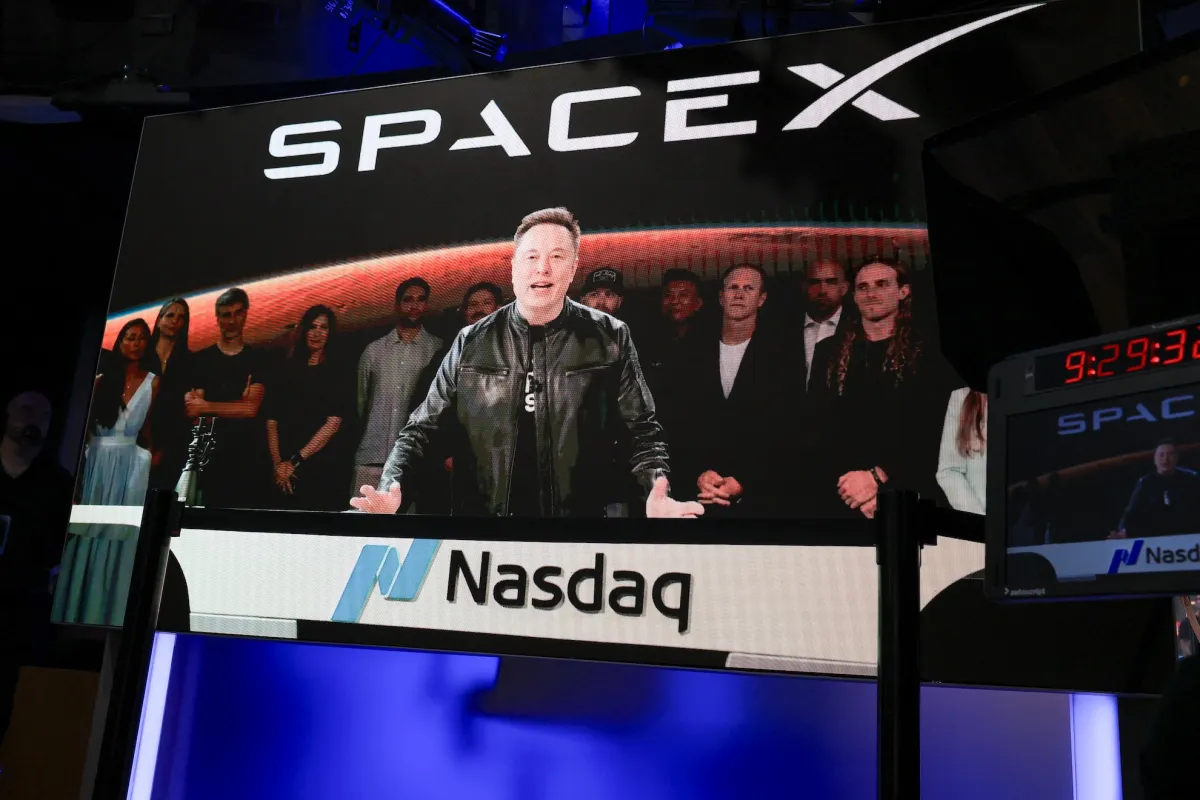A large screen displays a man speaking with a group behind him, SpaceX logo above, Nasdaq sign below, digital clock shows...